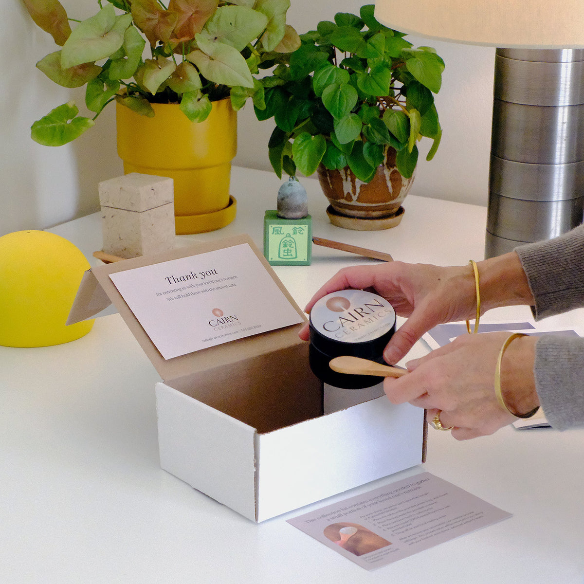 Hands opening a Cairn collection kit with a jar and small wooden spoon on a sunny table