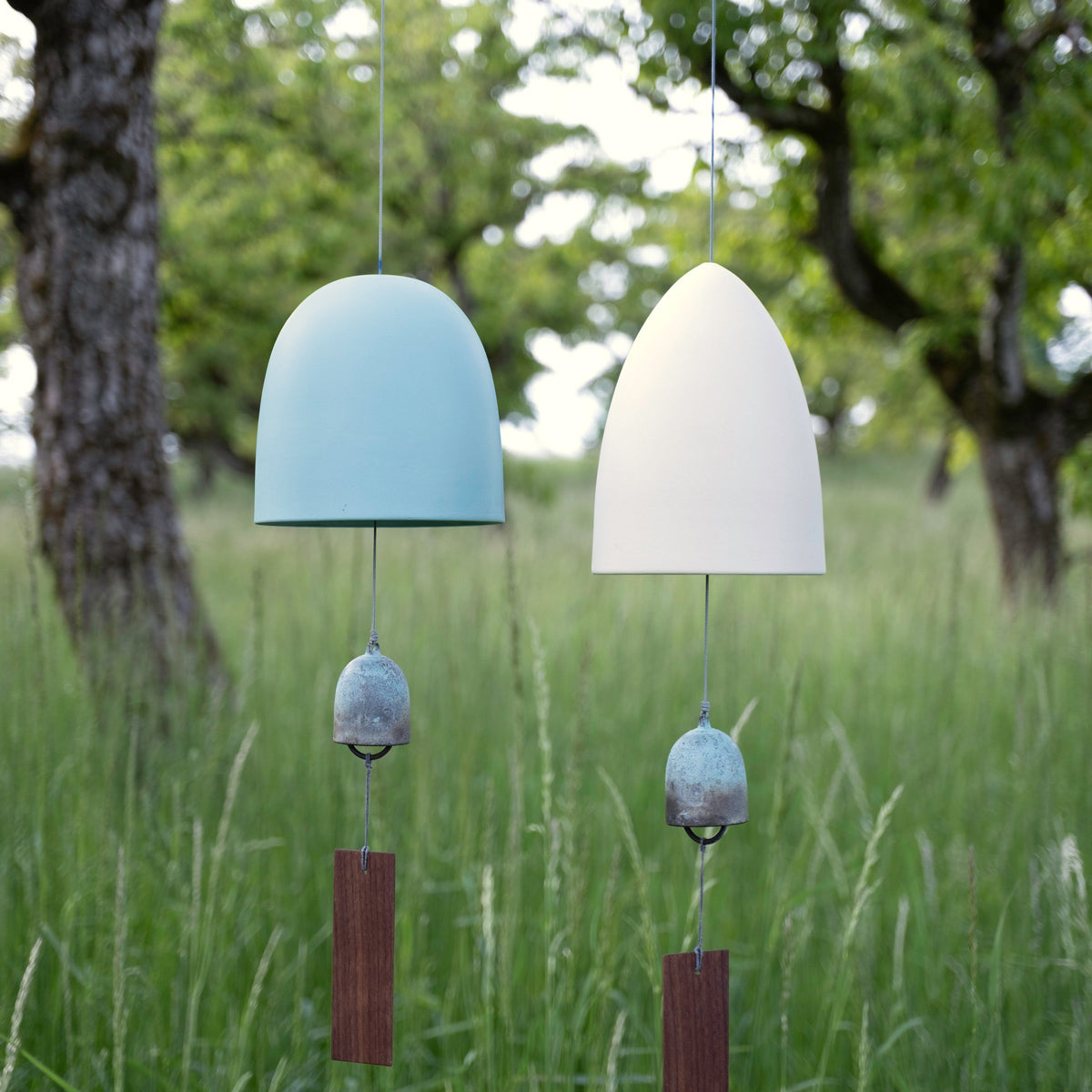 Two porcelain memorial wind chimes, pale blue and white, with bronze bells and walnut wind catchers, hanging in a grassy orchard
